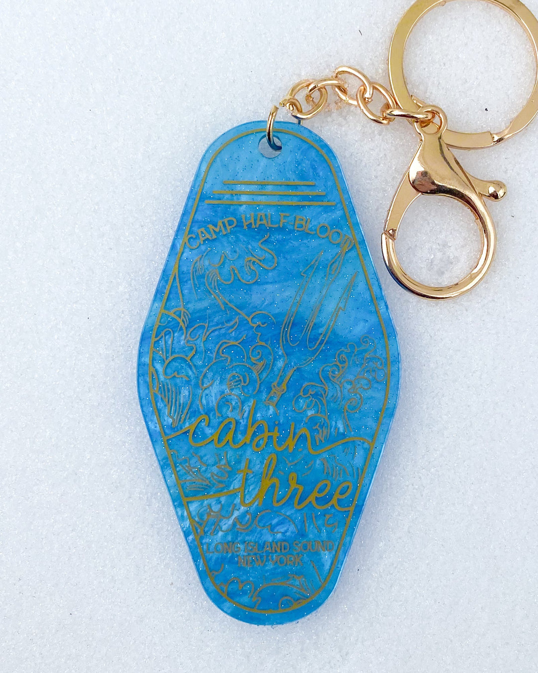 Demigod keychain featuring a blue background and gold detailing for a Cabin Three motel keychain with waves and a trident.
