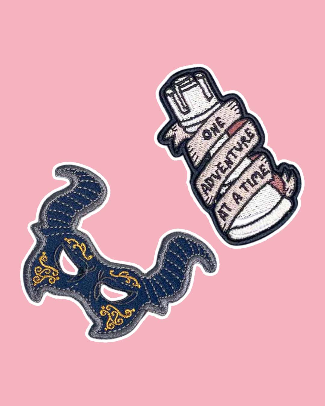 A Darker Shade of Magic patch set featuring a rook patch with a banner wrapped around it with 'One adventure at a time' on it and a masquerade mask in blue and gold for the other.