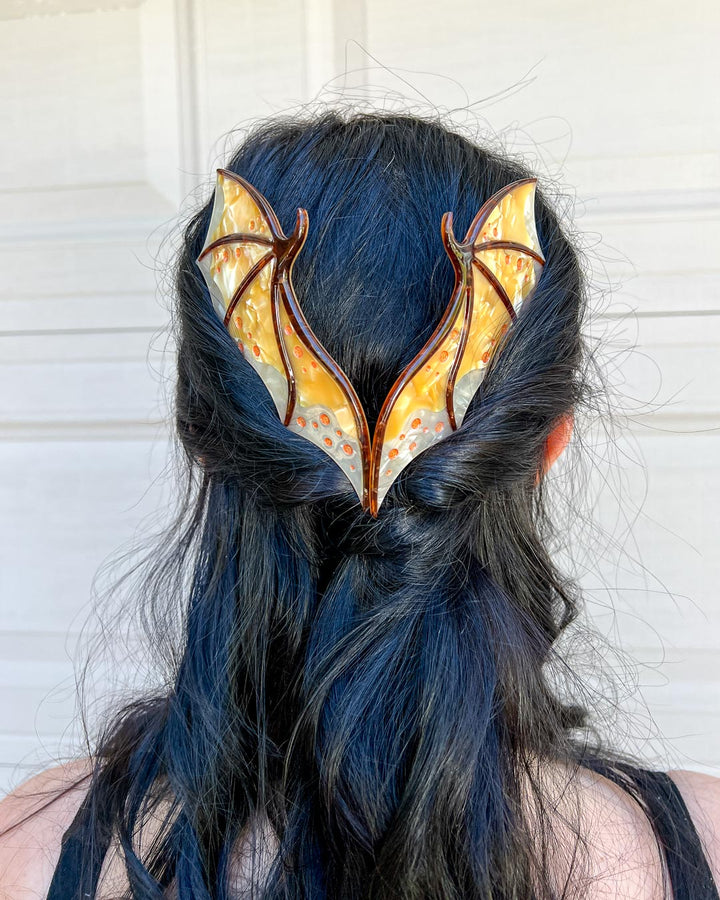 Fantasy hair combs featuring large combs in the shape of dragon wings in gold with engraved embellishments and a pearlescent color.