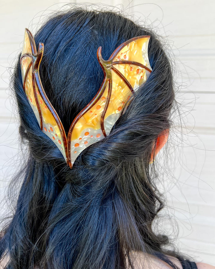 Hair combs for fantasy fans featuring two gold combs in the shape of dragon wings with engraved embellishments and a pearlescent gold color.