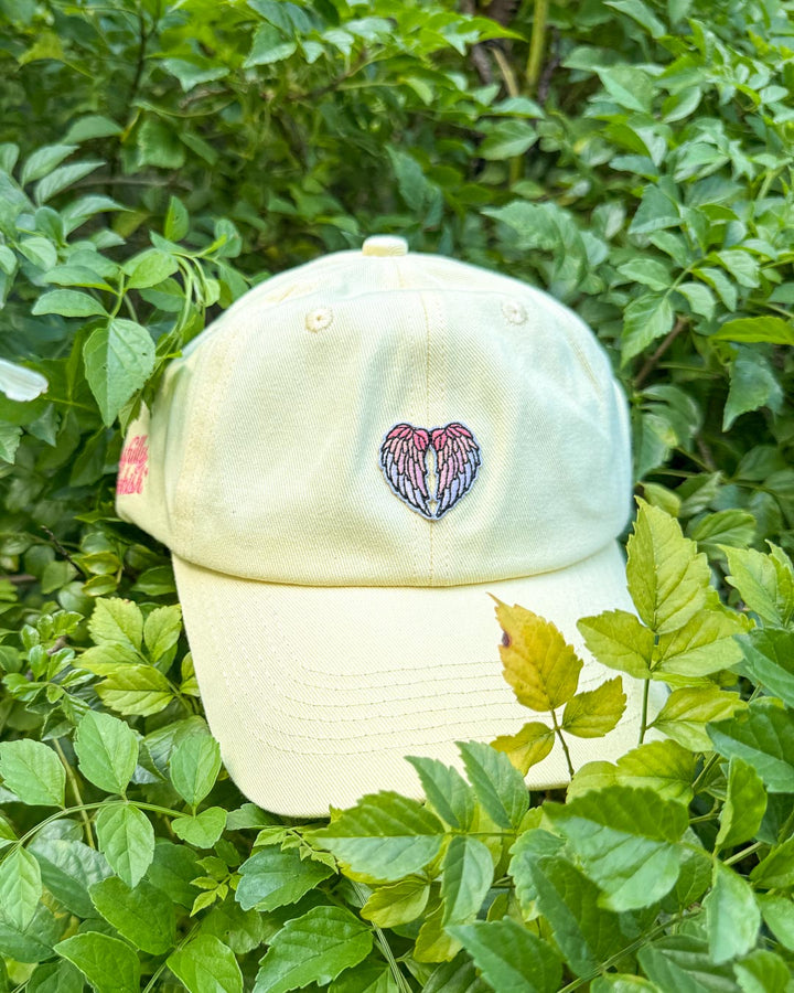 Light yellow romantasy hat featuring an angel wing design on a green leafy background.