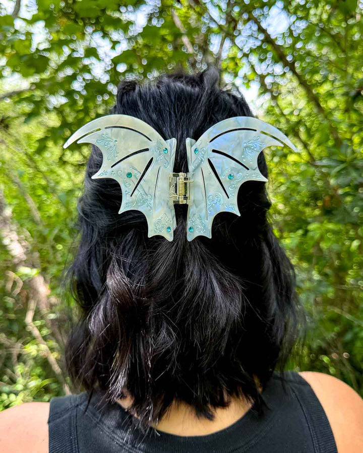 A pearlescent bookish hair clip in pastel blue with blue gem embellishments in the shape of bat wings inspired by ACOTAR.