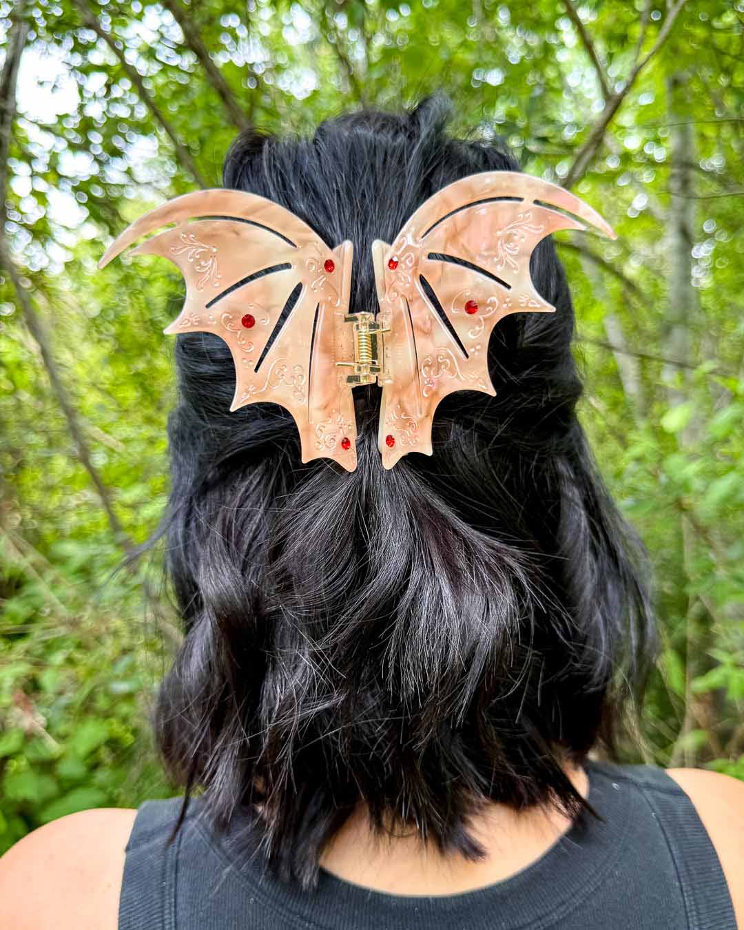 A pearlescent bookish hair clip in pastel red with red gem embellishments in the shape of bat wings inspired by ACOTAR.
