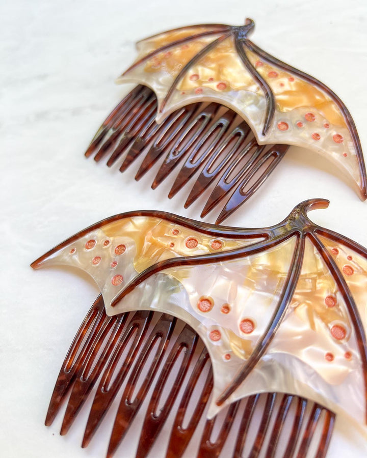 Close up of a fantasy hair accessory in the shape of dragon wings, featuring two combs in a gold pearlescent color and engraved details.