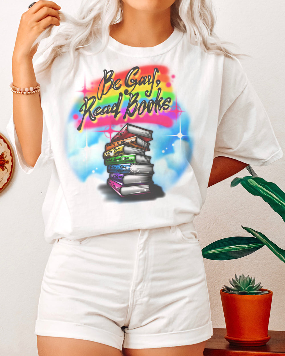 Bookish shirt featuring an airbrushed design of a rainbow stack of LGBTQ+ books and the phrase 'Be gay, read books.'