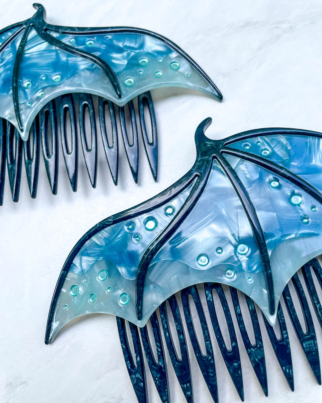 Close up of a fantasy hair accessory in the shape of dragon wings, featuring two combs in a blue pearlescent color and engraved details.