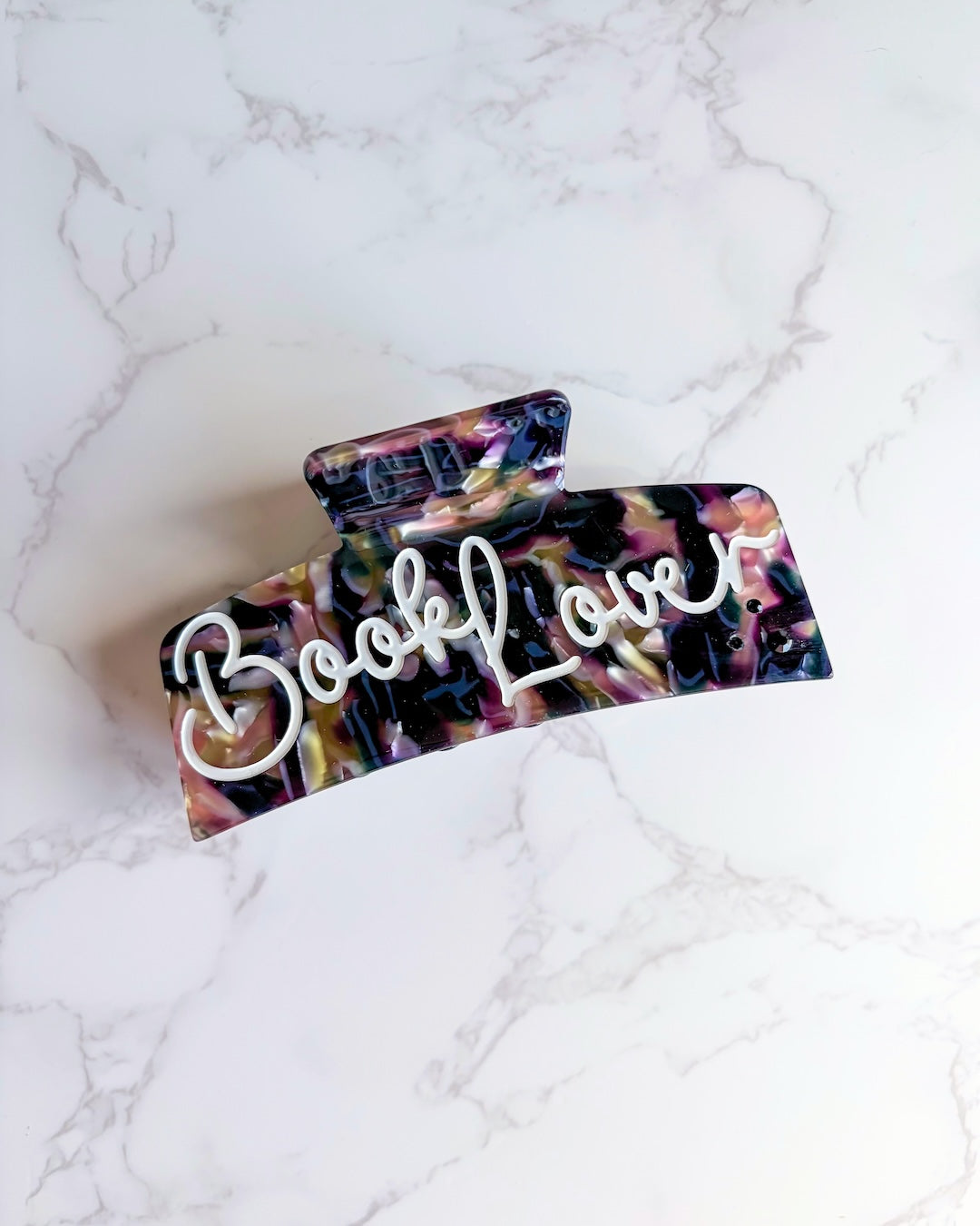 Flat lay of the book lover hair claw clip in a classic rectangular claw clip shape with "Book Lover" etched in white script font over a dark, multi-colored marbled base on a white marble background.