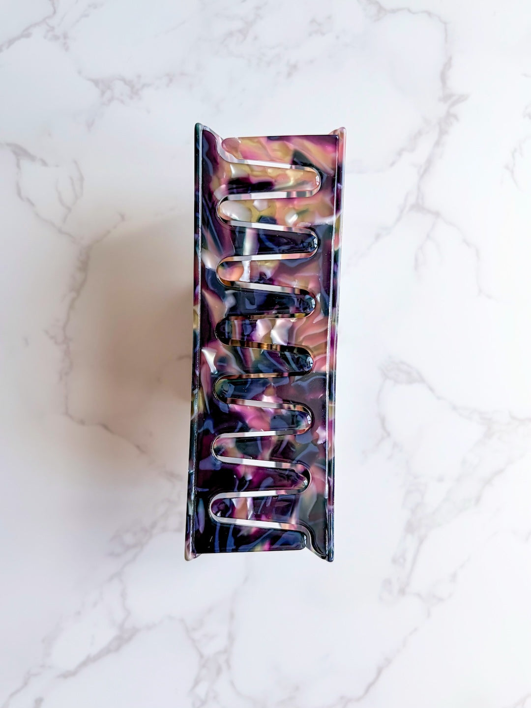 Bottom of a hair clip for book lovers featuring the wide claws in dark, multi-colored marbled colors on a white marbled countertop background.
