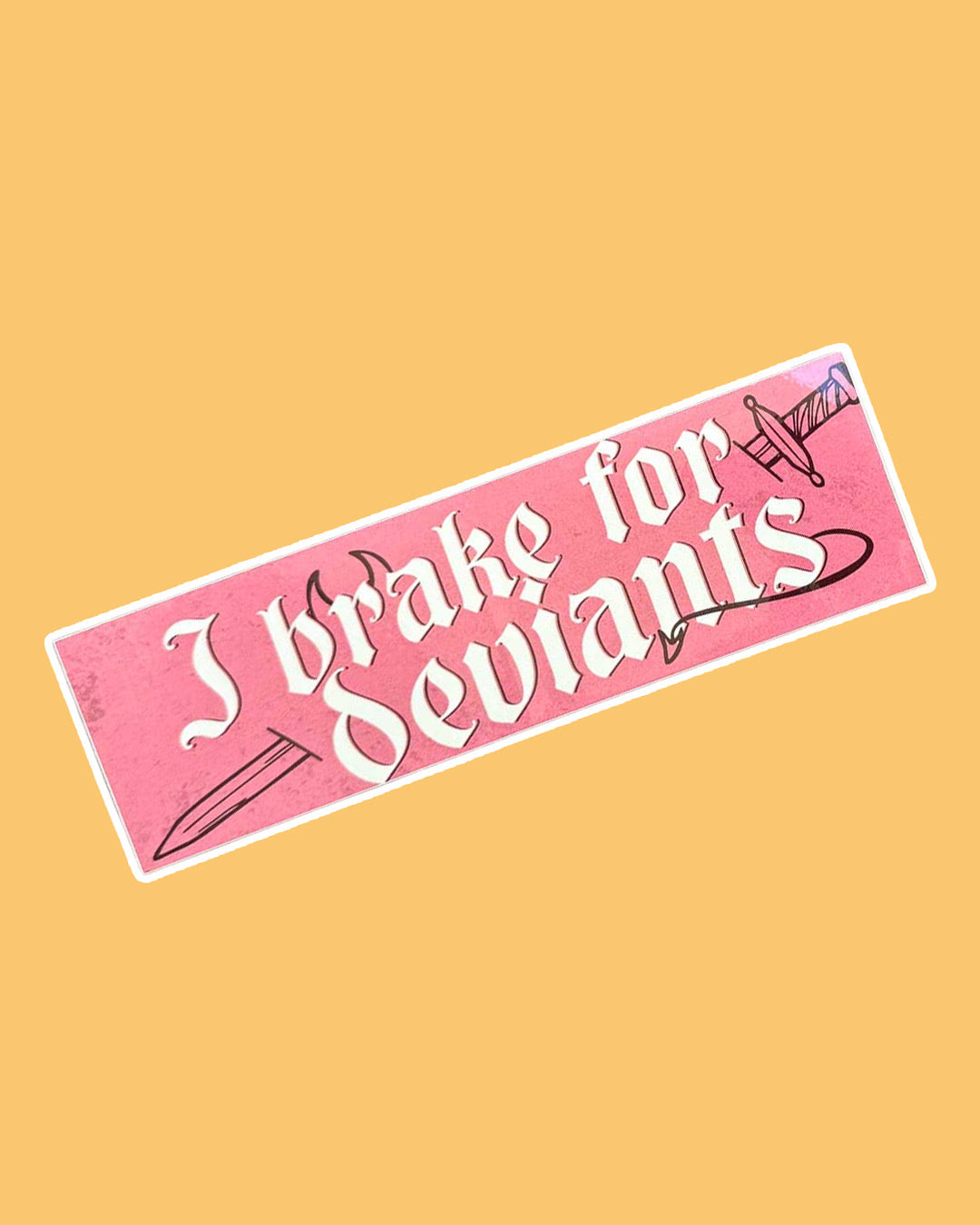 Pink bumper sticker on yellow background featuring the phrase "I brake for deviants" in a medieval-esque font with devil horns sprouting above "brake," a devil's tail curling off the 's' in "deviants," and a sword running through the words.