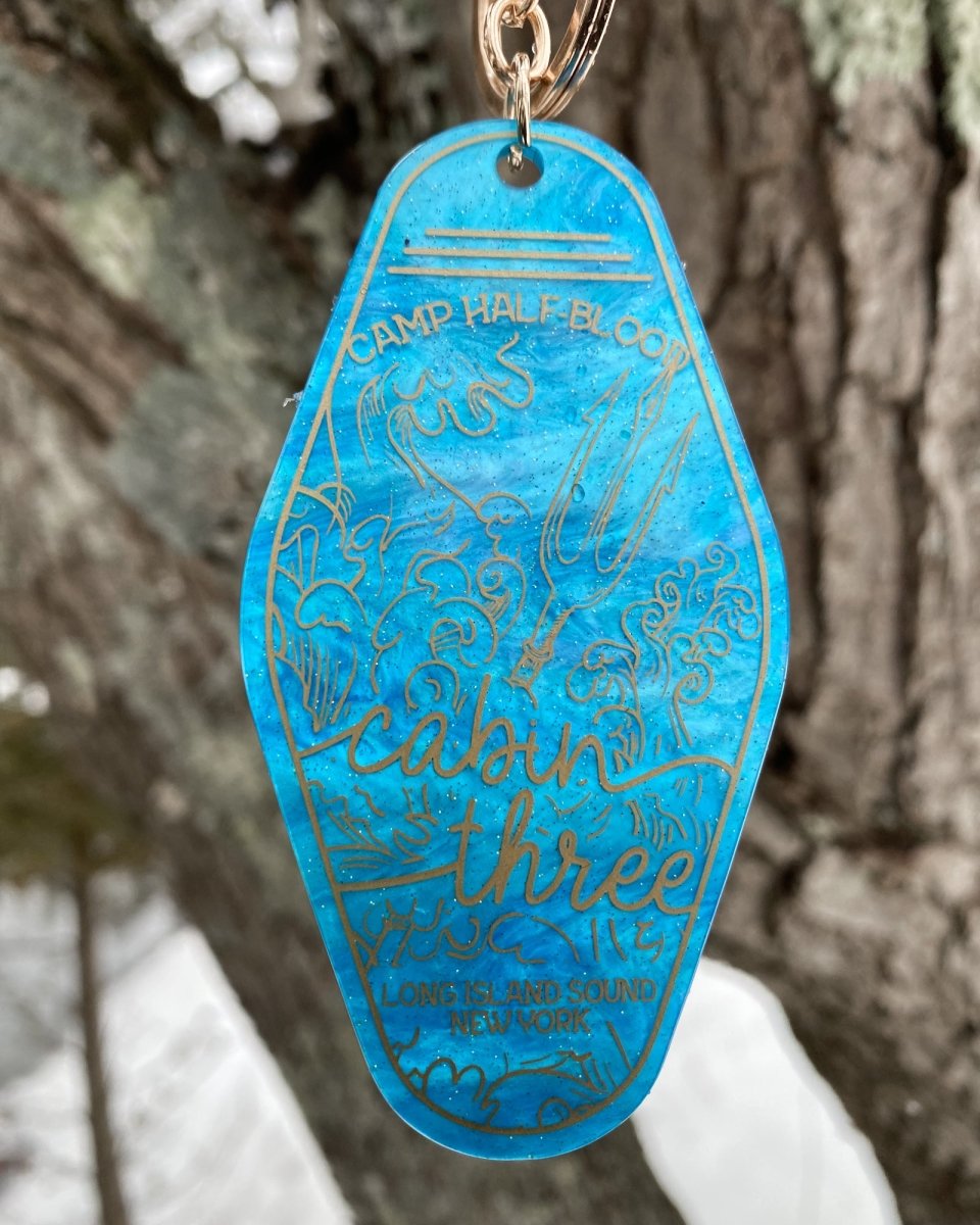 PJO keychain for Camp Half-Blood's Cabin Three featuring a blue background and gold illustrations of waves and a trident.