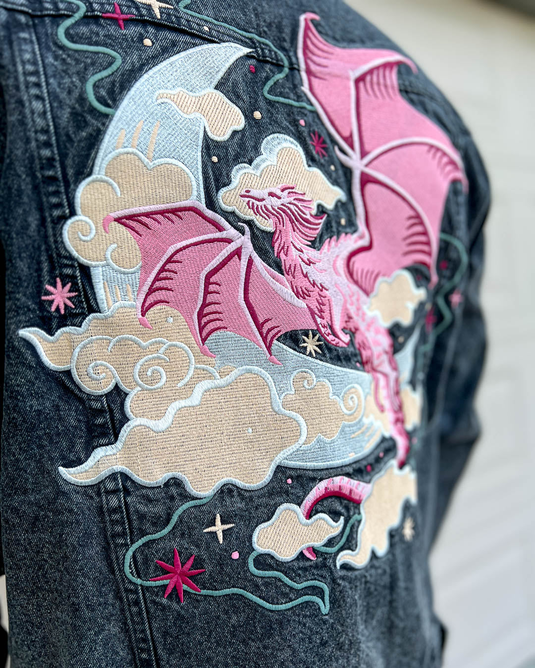 Celestial dragon oversized jean jacket with a vibrant embroidered design of a pink flying dragon over a crescent moon amongst stars and clouds.