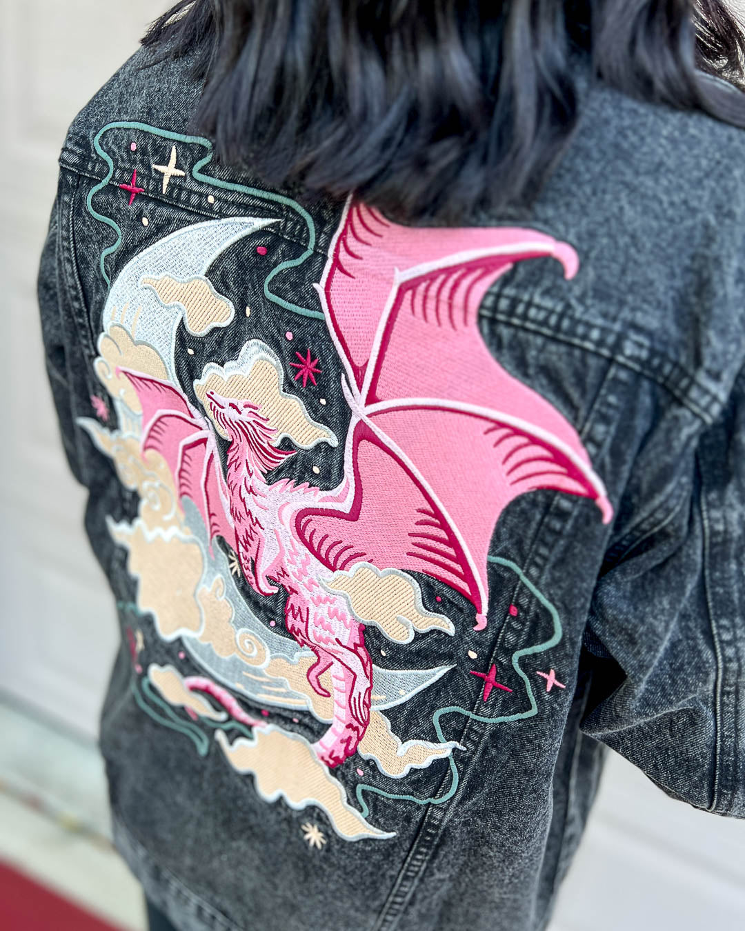 Celestial dragon jean jacket featuring an oversized fit, an embroidered pink dragon, a crescent moon, clouds, stars, and a colorful sleeve detail.