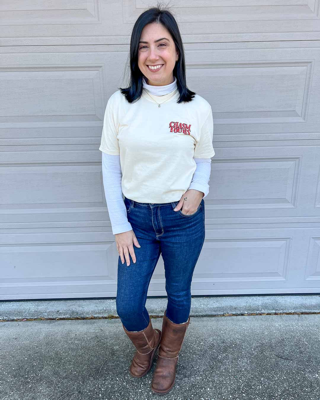 Woman with black hair, in jeans, brown boots, and a longsleeve white shirt wearing an ivory "Chasm Tours" shirt over it, inspired by Kate Golden&