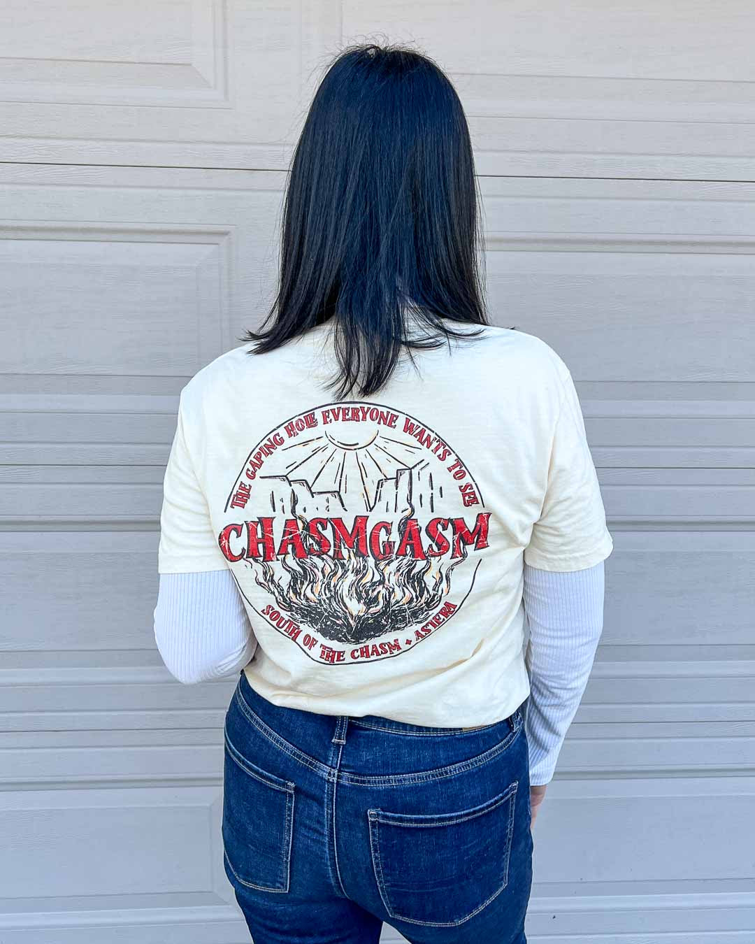 Back of the Chasm Tours shirt featuring a circular logo of a sun shining down on a chasm with fire coming up from the bottom that reads "Chasmgasm" across the middle and the text curving around the border on the top reading "The gaping hole everyone wants to see" and "South of the chasm, Astera" curving along the bottom.