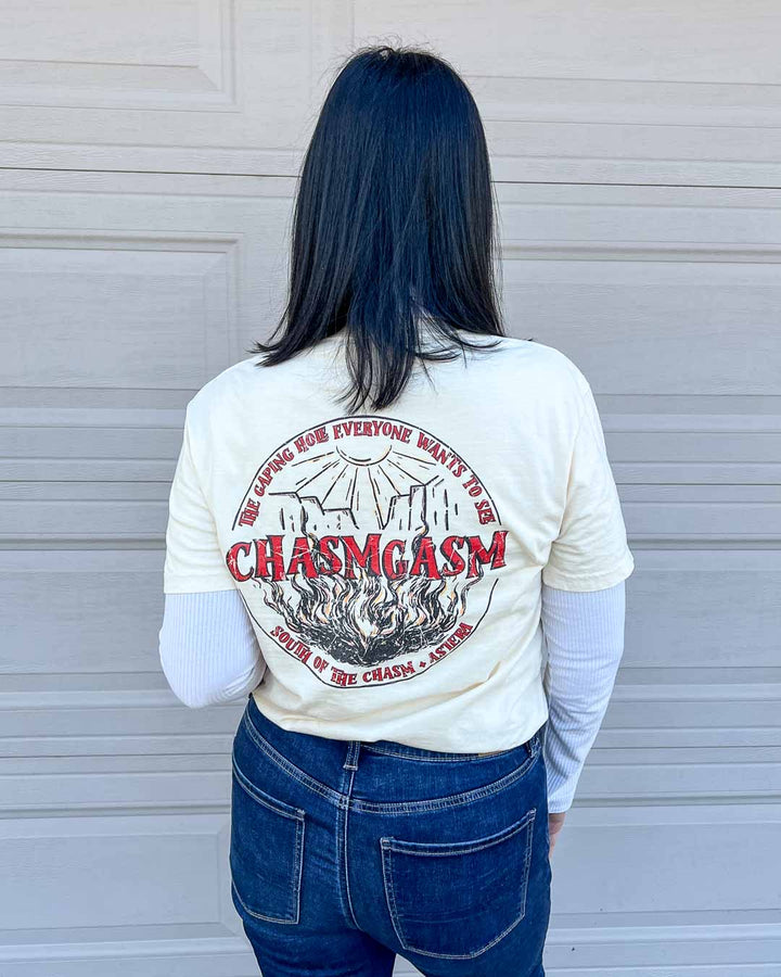 Back of the Chasm Tours shirt featuring a circular logo of a sun shining down on a chasm with fire coming up from the bottom that reads "Chasmgasm" across the middle and the text curving around the border on the top reading "The gaping hole everyone wants to see" and "South of the chasm, Astera" curving along the bottom.