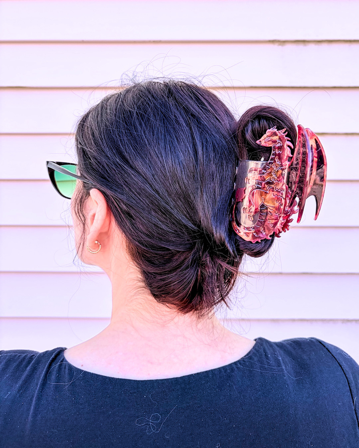 Woman with dark hair with her head turned to the side, showing off a fantasy hair claw clip for book lovers, featuring an iridescent red dragon holding her hair up against a pinkish background.