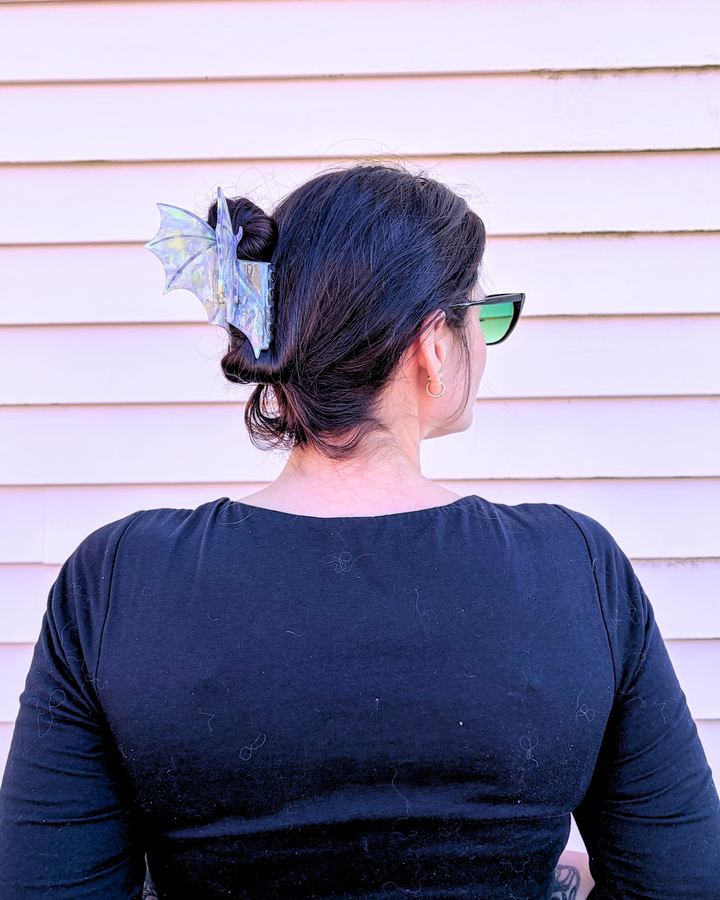 Woman wearing a purple hair claw clip for fantasy readers, featuring dragon wings spread wide with an iridescent shine, all on a pinkish background.