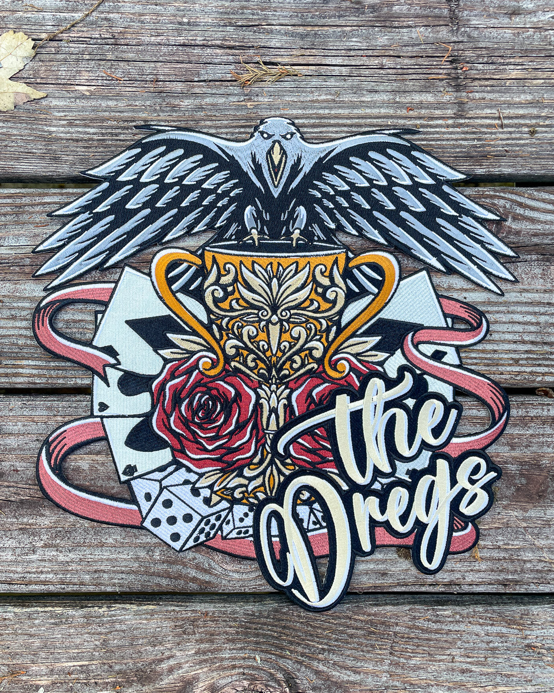 Flat lay of the Dregs patch for Six of Crows fans featuring a crow, a cup, roses, playing cards, and dice.