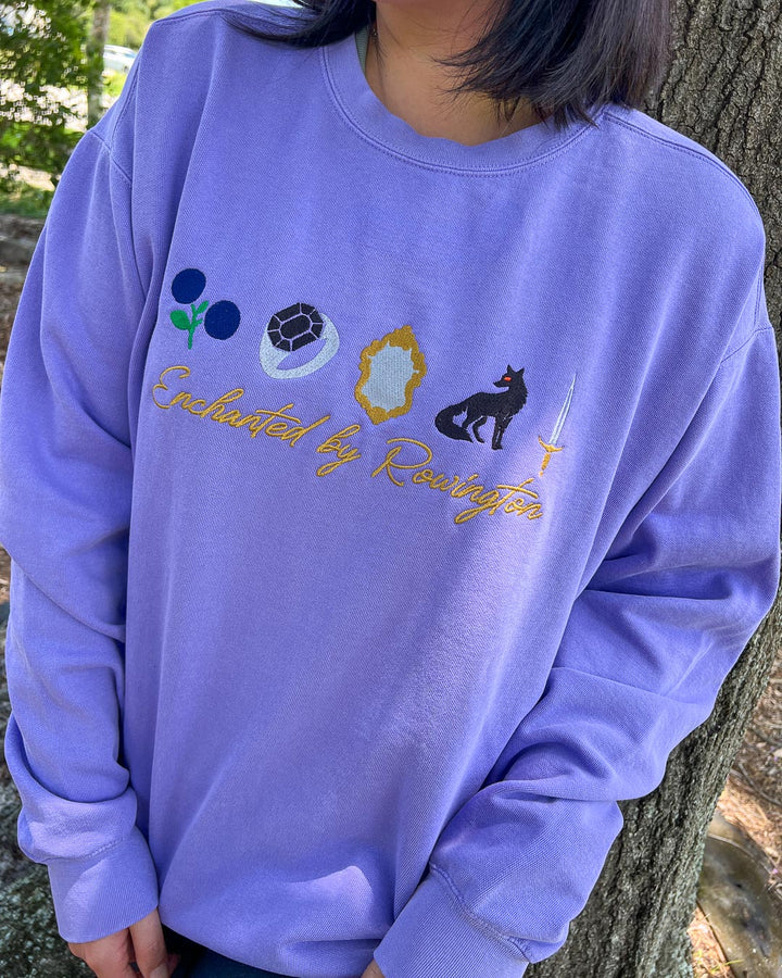 Person wearing a purple sweatshirt with embroidered designs of berries, a ring, a mirror, a fox, and a sword, with the text &