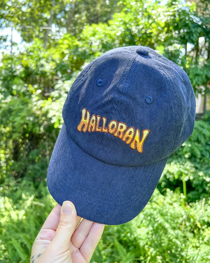Person holding the If Not For My Baby baseball cap featuring a yellow and orange logo for Halloran on navy blue corduroy.