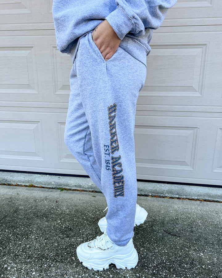 Woman modeling the Harker Academy sweatpants, featuring "Harker Academy" written in all caps and traced in gold edging down the left pant leg and "EST 1845" beneath it.