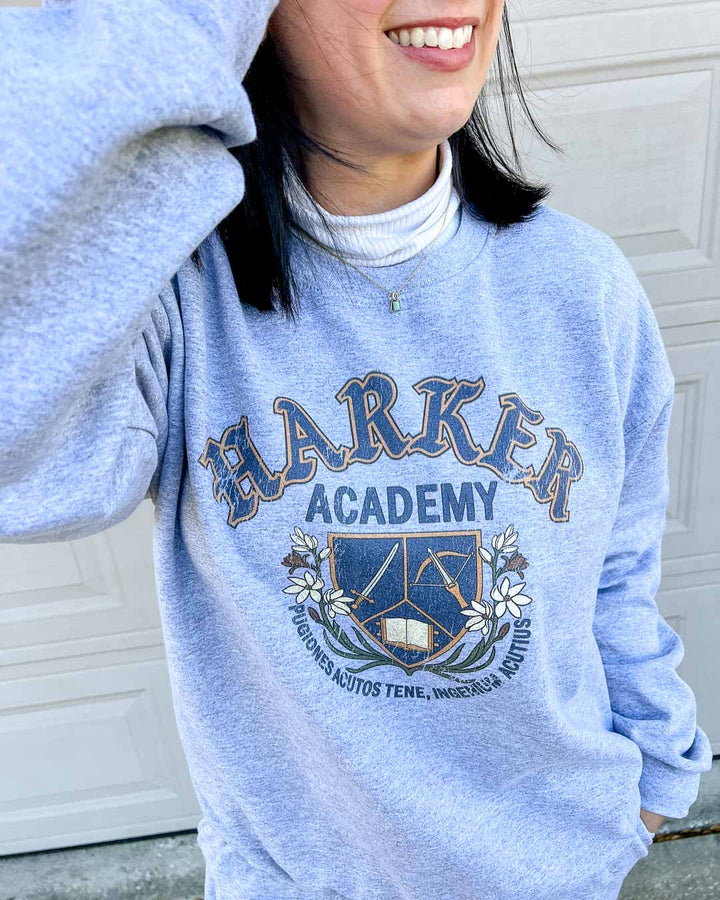 Close up of the Half City gray crew neck sweatshirt featuring a distressed design reading "Harker Academy" and a logo of a sword, crossbow, and open book beneath it.