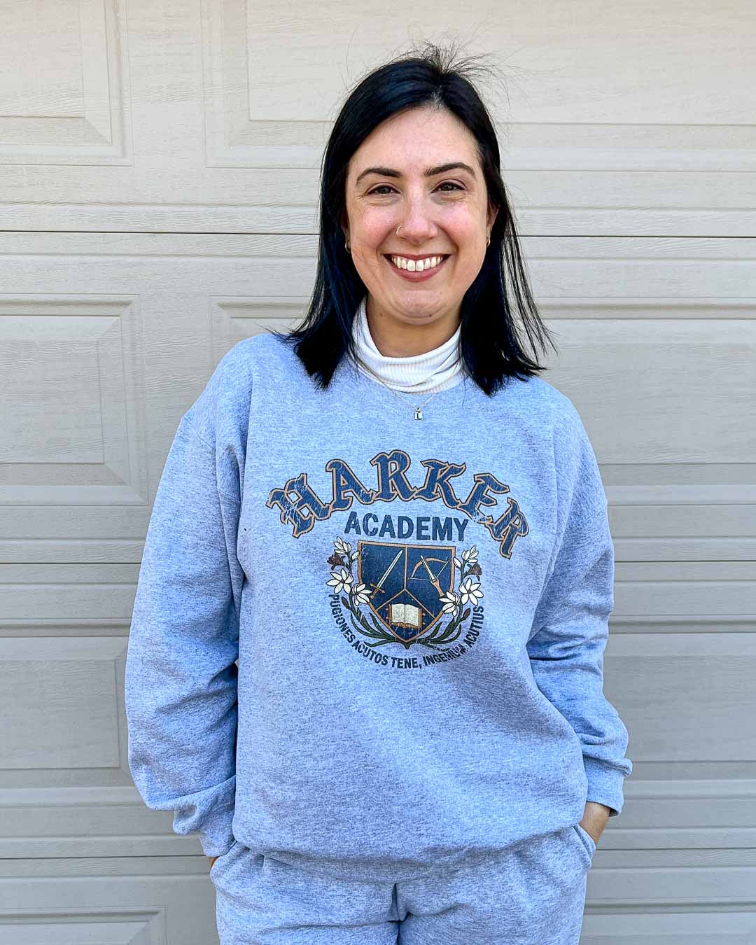 Model wearing a fantasy sweatshirt featuring a crest for Harker Academy, a fictional magic school from Half City, designed in a shield with a sword, crossbow, and open book, and a motto in Latin curling beneath it.