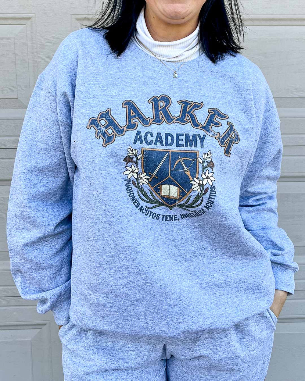 Women wearing a gray Harker Academy sweatshirt featuring a logo for the magic school from Half City by Kate Golden featuring a shield bracketed by flora and branches with a sword, a crossbow, and an open book with a motto in Latin beneath it.