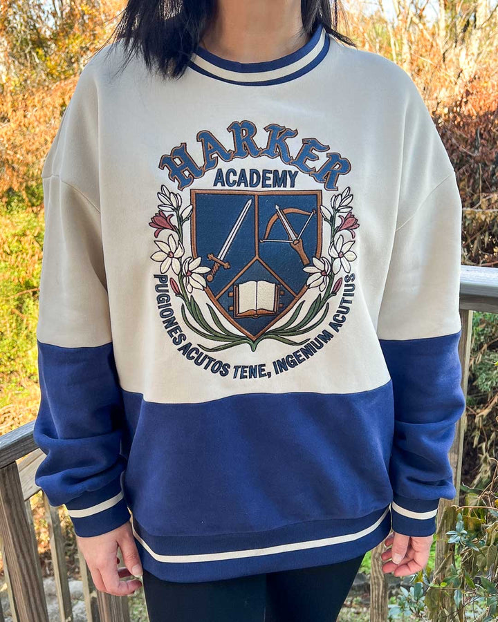 Model wearing a fantasy blue and white sweatshirt featuring a crest for Harker Academy, a fictional magic school from Half City, designed in a shield with a sword, crossbow, and open book, and a motto in Latin curling beneath it.