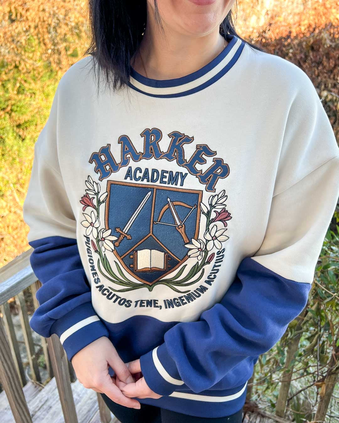 Women wearing a dual-tone blue and white Harker Academy sweatshirt featuring a logo for the magic school from Half City by Kate Golden featuring a shield bracketed by flora and branches with a sword, a crossbow, and an open book with a motto in Latin beneath it.