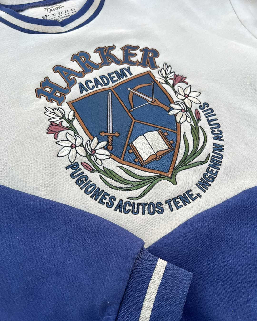 Close up of the Half City gray crew neck sweatshirt featuring an embroidered design reading "Harker Academy" and a logo of a sword, crossbow, and open book beneath it.