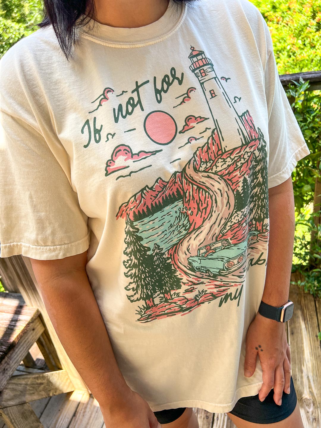 Kate Golden t-shirt featuring an illustrated landscape design with the sun setting over a lighthouse and two people driving in a blue, vintage car with &