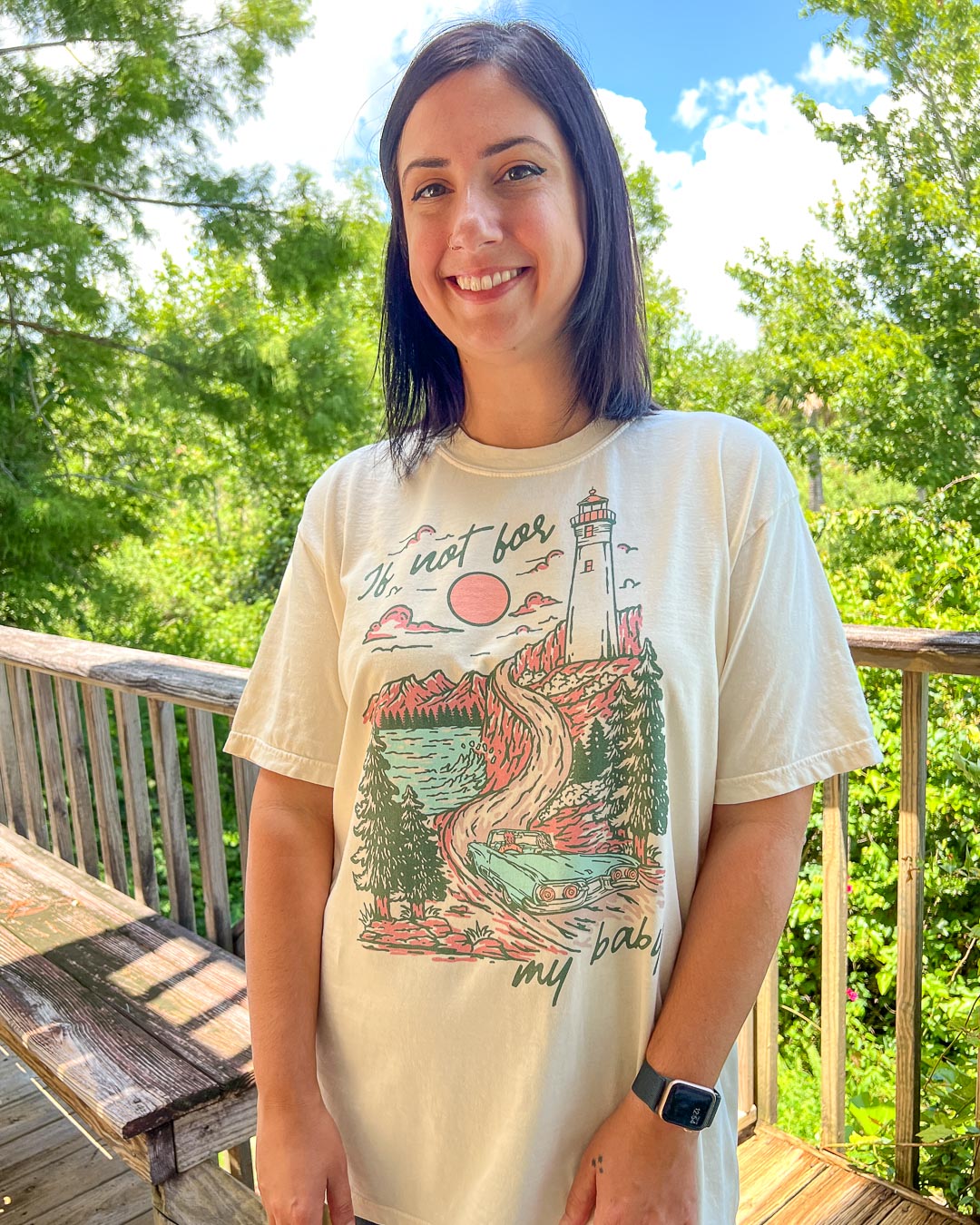Woman wearing a Kate Golden shirt for If Not For My Baby fan featuring an illustrated landscape of a lighthouse and a blue vintage car.