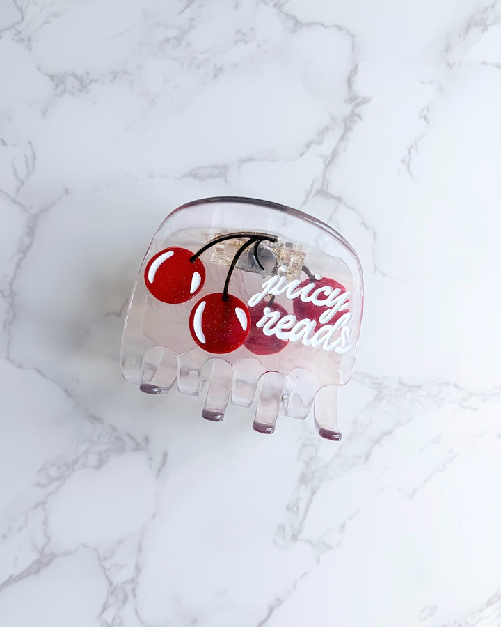 Clear claw clip featuring two cherries and "juicy reads" written in white font on it on a white, marbled background.