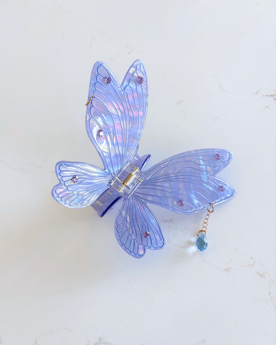 Pale purple fairy wing hair claw clip for fantasy readers featuring jewel embellishments on the wings and a  dangling gem against a white marble background.