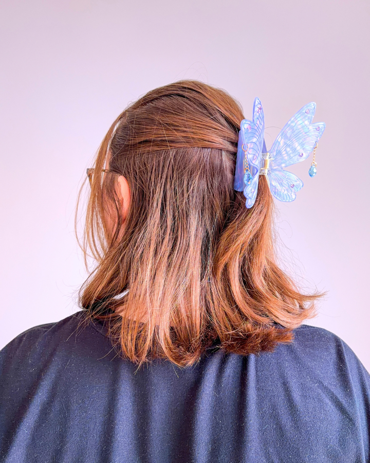 Woman with short brown hair with her head turned to the side, hair half up half down with a fantasy hair clip in her hair, featuring a fairy wing design in lavender purple with blue undertones, etching details, gem embellishments, and two dangling jewels.