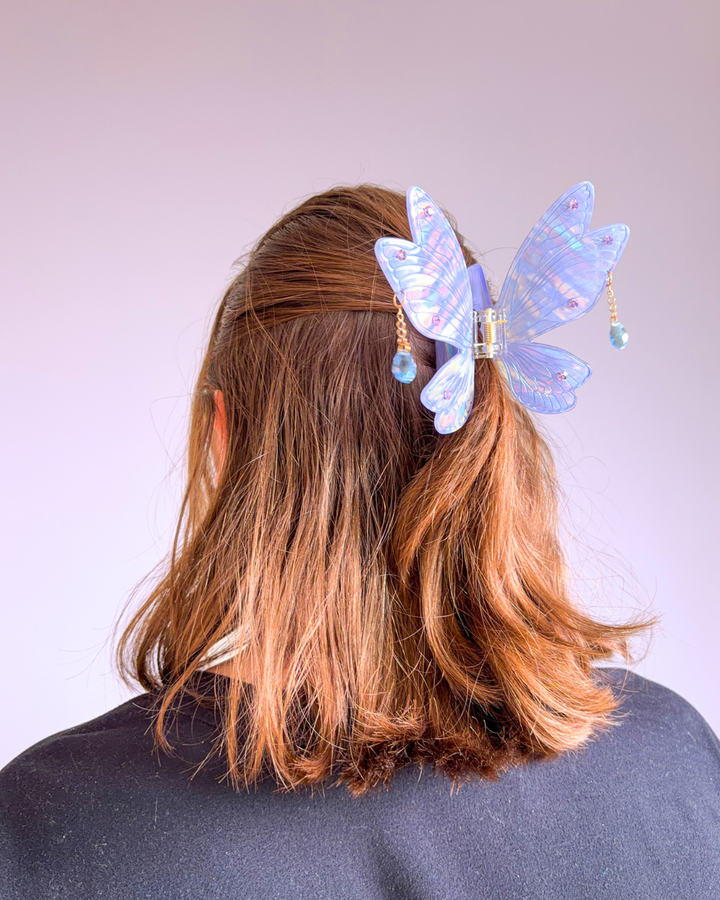 Woman wearing her hair in a half up half down style clipped with a lavender claw clip in the shape of fairy wings with dangling gems off the wings.