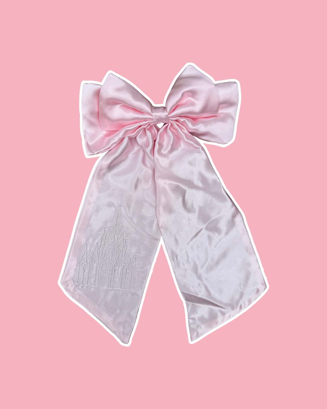 Pink satin hair bow featuring an embroidered design of a magical castle on the left tail on a pink background.