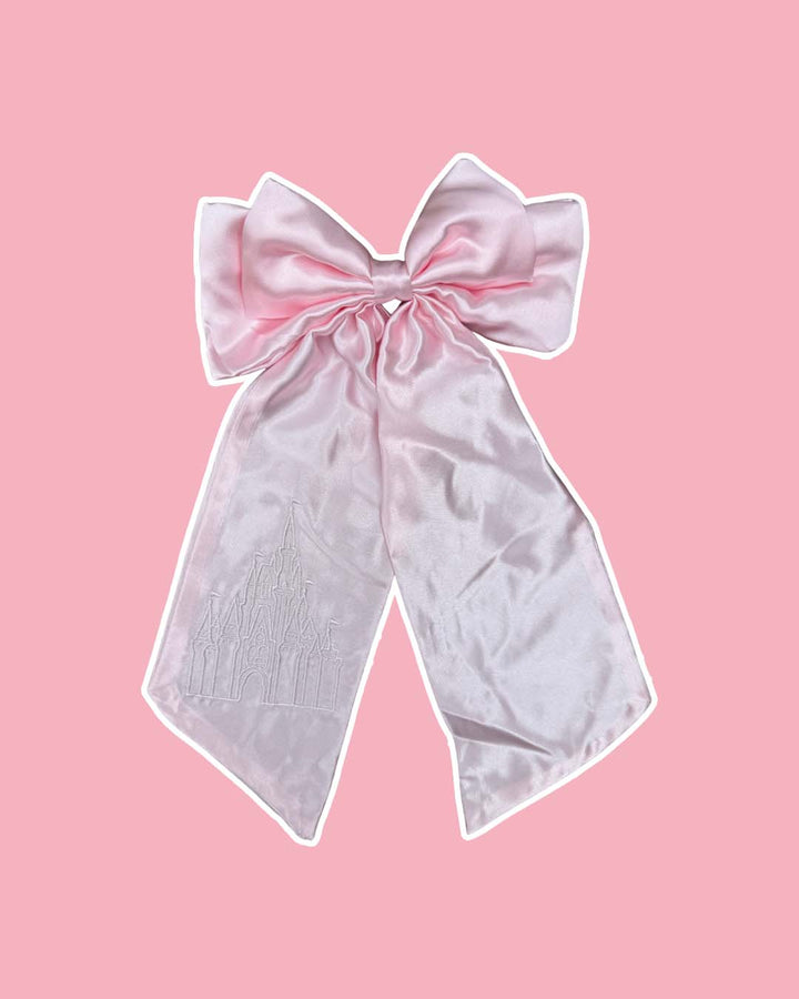 Pink satin hair bow featuring an embroidered design of a magical castle on the left tail on a pink background.