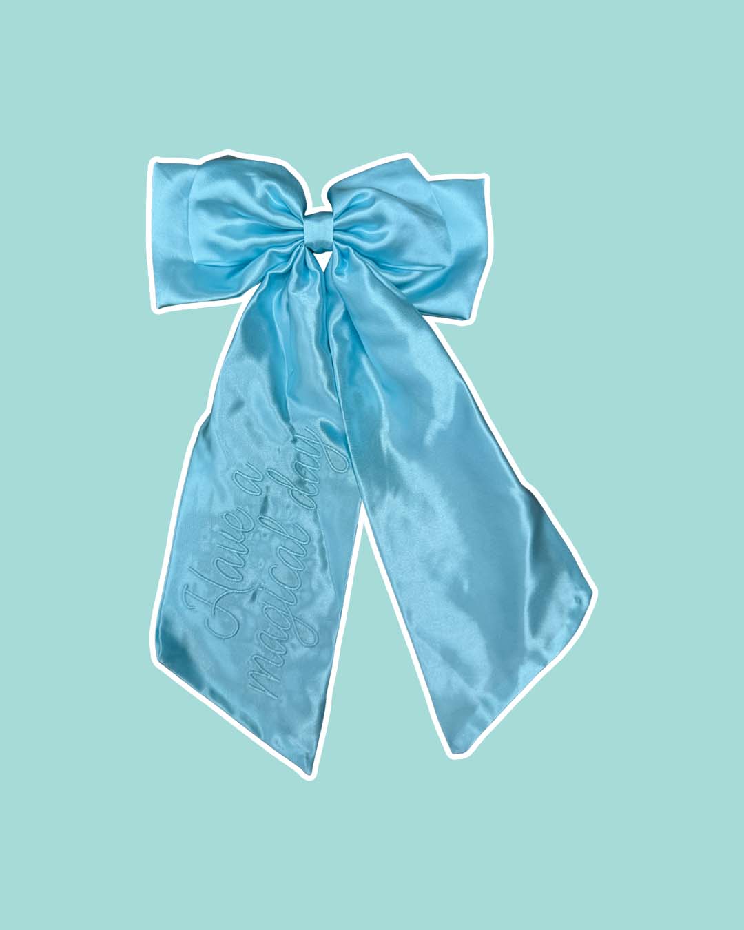 Blue satin hair bow reading 'Have a magical day' in embroidered script text on a light blue background.
