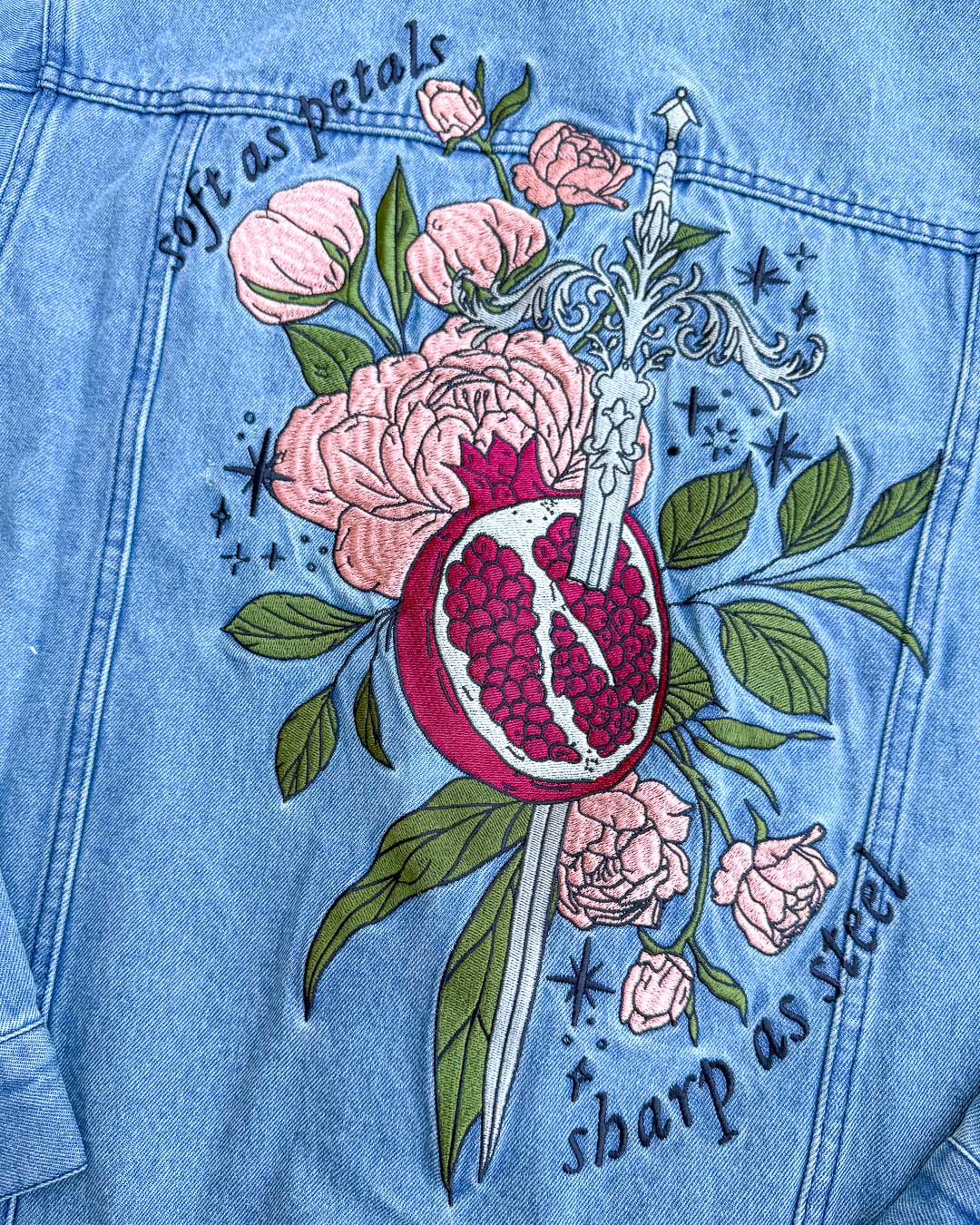 Persephone jacket designed with an embroidered illustrated sword spearing through a pomegranate over leaves, roses, and stars, with the quote &