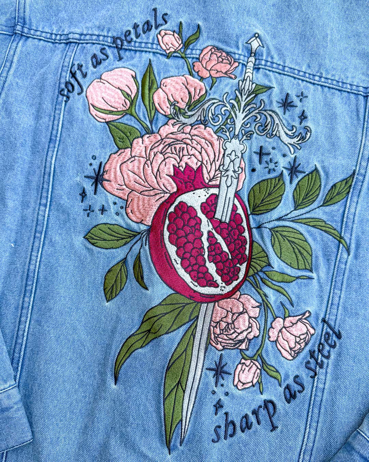 Persephone jacket designed with an embroidered illustrated sword spearing through a pomegranate over leaves, roses, and stars, with the quote &