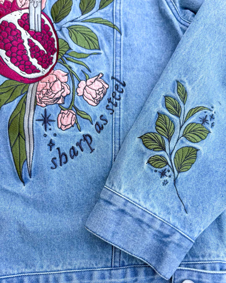 Close up of the Persephone jean jacket highlighting the leaves and stars on the sleeve and the embroidered back of the jacket featuring the sword spearing a pomegranate, leaves, roses, and the phrase &