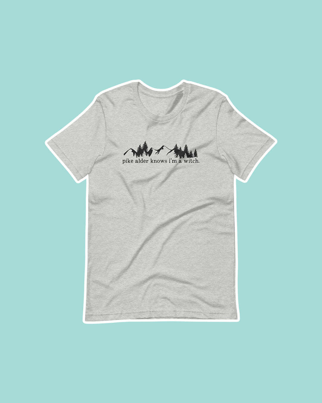 Wild is the Witch shirt featuring the phrase 'Pike Alder knows I'm a witch' embroidered beneath an illustrated silhouette of mountains and pine trees.