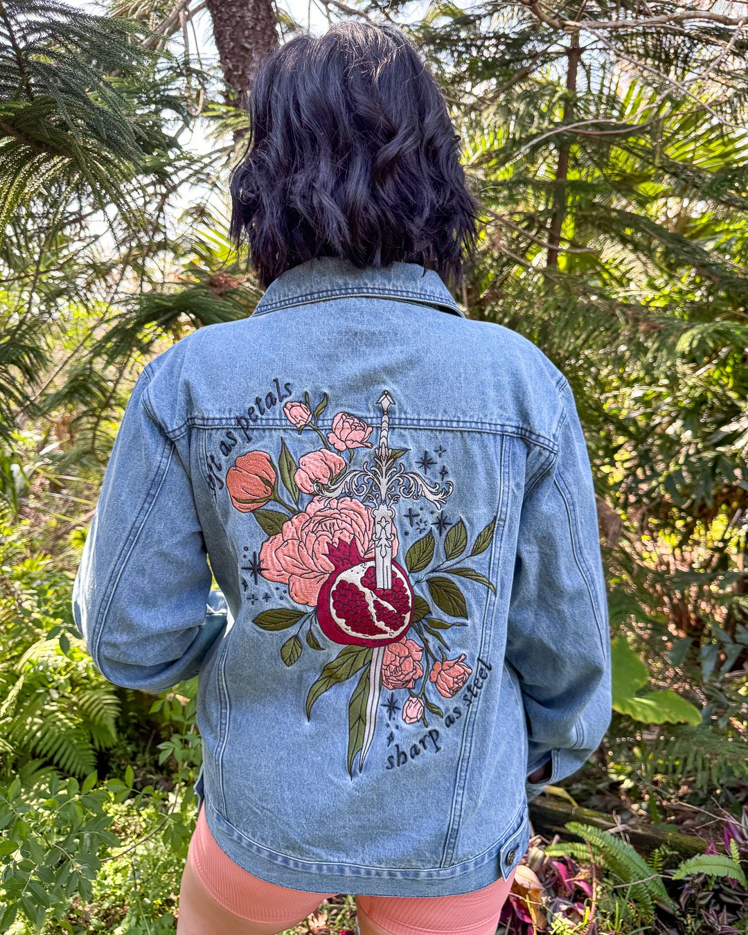 Embroidered fantasy outerwear for mythology fans featuring a design on the back of a sword going through a pomegranate on the backdrop of leaves and roses with the phrase, &