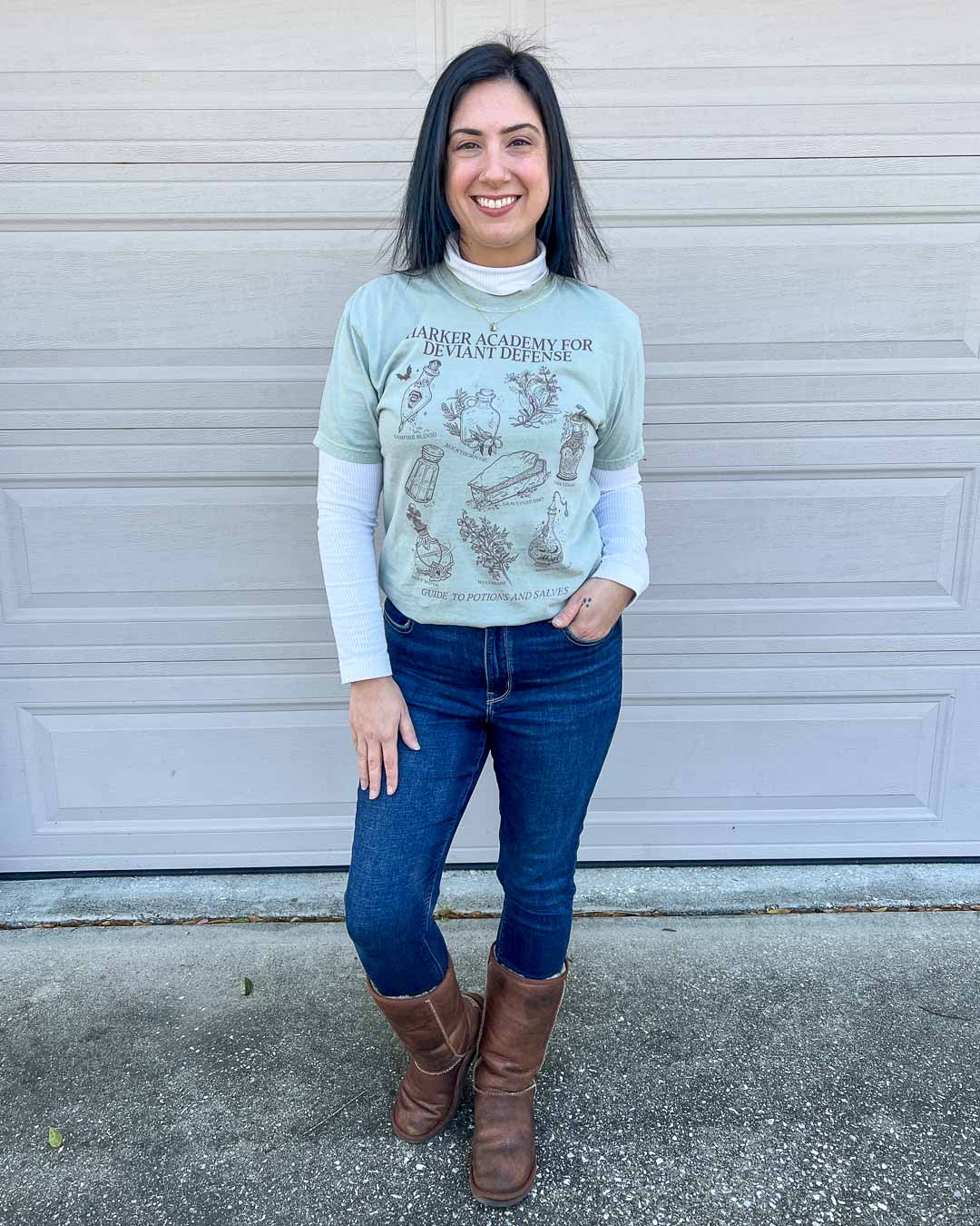 Woman wearing jeans, brown boots, a long sleeved white shirt, and the Potions &amp; Salves shirt over it inspired by Half City by Kate Golden.