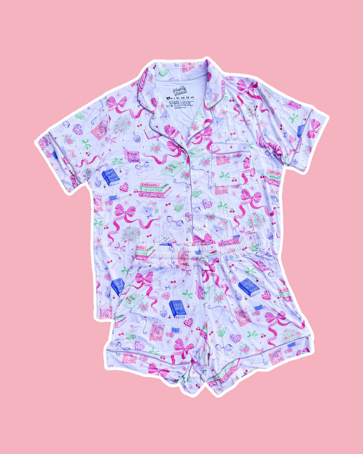 Romance tropes pjs set with collared button down shirt, a pocket, and a stretchy waistband with a printed design featuring illustrated pink bows and stacks of books with different tropes on them.