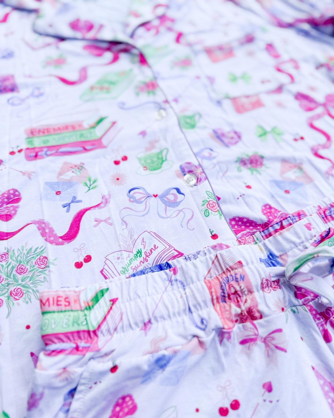 Close up of the romance trope pajamas set featuring illustrated bows, teacups, and books listed with tropes like enemies to lovers, grump sunshine, forbidden love, and more.