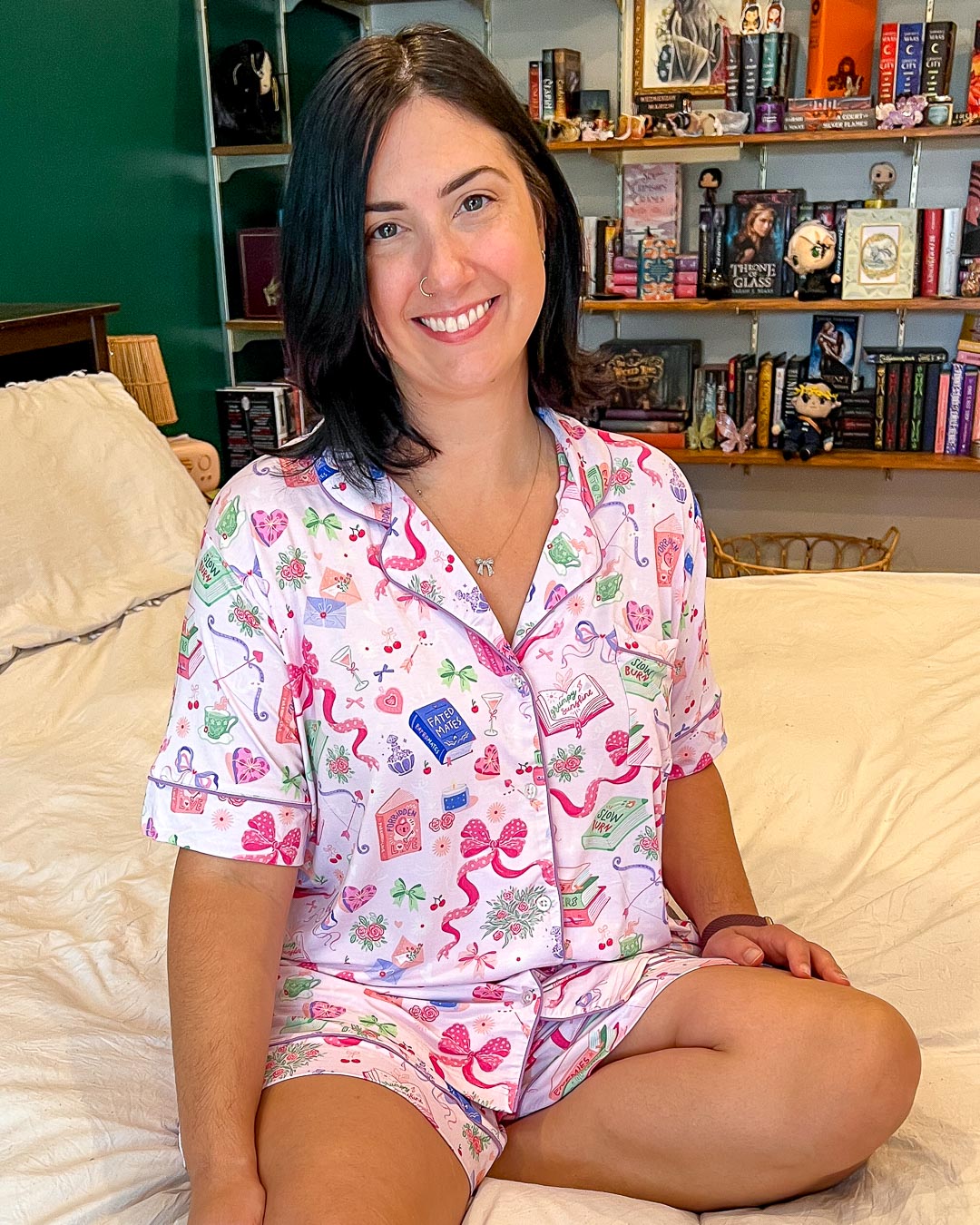 Model wearing the trope pjs set for romance readers covered in illustrated designs of bows, teacups, flowers, and books with different tropes on them like fated mates, slow burn, grump sunshine, and more.
