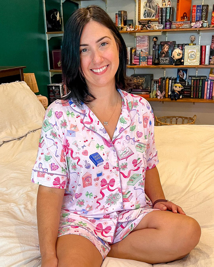 Model wearing the trope pjs set for romance readers covered in illustrated designs of bows, teacups, flowers, and books with different tropes on them like fated mates, slow burn, grump sunshine, and more.